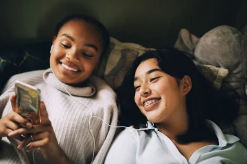 Op de afbeelding zijn twee jonge meiden te zien die lachend samen naar een mobiel kijken.